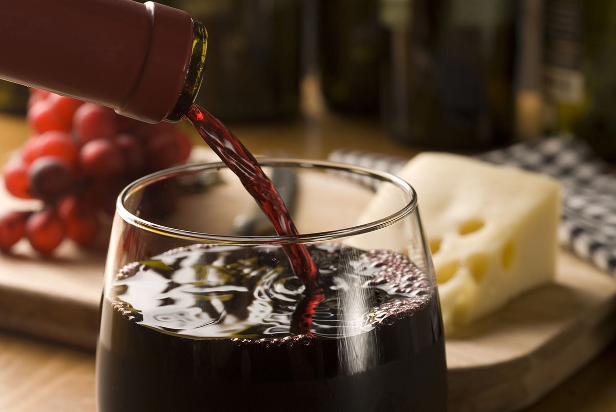 Merlot wine poured into a glass, with cheese and fruits in the background.